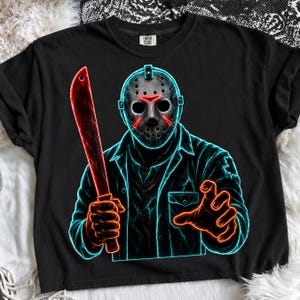 May include: A black t-shirt featuring a neon-style graphic of a hockey-masked figure holding a machete. The design uses vibrant blue outlines and red accents to depict the character, who wears a collared shirt with a chest pocket and distressed details.