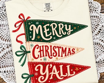 May include: A cream-colored t-shirt featuring a graphic of three stacked pennants. The top pennant is green with the text MERRY, the middle is cream with CHRISTMAS, and the bottom is red with Y'ALL. Each pennant has a ribbon and star accents.