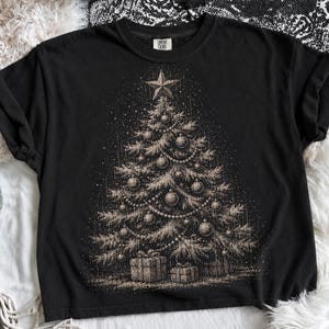 May include: A black short-sleeve t-shirt featuring a detailed, light-colored stippled illustration of a decorated Christmas tree. The tree is adorned with ornaments, garlands, and a star topper, with wrapped gift boxes placed at the base.
