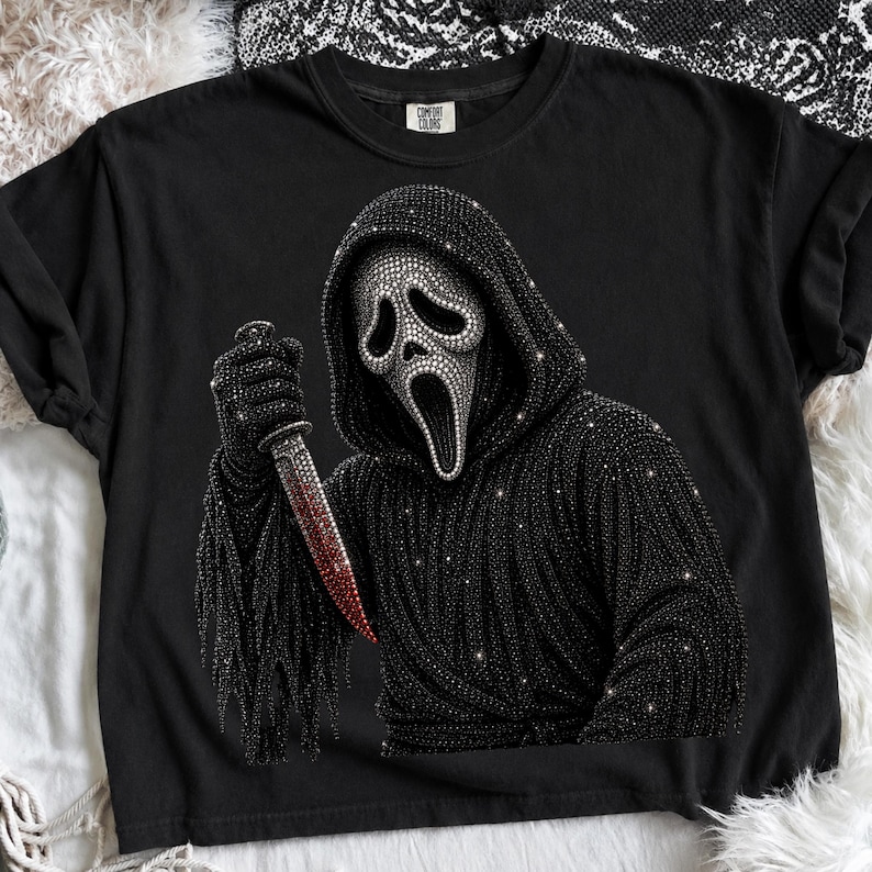 May include: A black short-sleeved t-shirt featuring a graphic of a hooded figure with a white mask, covered in sparkling black rhinestones. The figure holds a knife with a red rhinestone blade. The shirt has a Comfort Colors brand label.