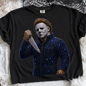 May include: A black t-shirt featuring a graphic of a character wearing a mask and a dark jumpsuit, holding a knife. The design is embellished with numerous small, sparkling rhinestones that create a shimmering effect across the mask, knife, and jumpsuit.