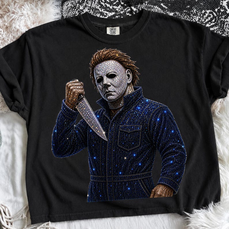 May include: A black t-shirt featuring a graphic of a character wearing a mask and a dark jumpsuit, holding a knife. The design is embellished with numerous small, sparkling rhinestones that create a shimmering effect across the mask, knife, and jumpsuit.