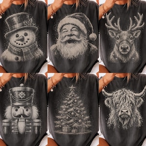 May include: A collection of six charcoal gray t-shirts, each featuring a distinct white graphic print. The designs include a snowman, a laughing Santa Claus, a reindeer, a nutcracker, a decorated Christmas tree, and a shaggy Highland cow.