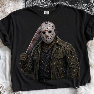 May include: A black t-shirt featuring a graphic of a hockey mask-wearing figure holding a baseball bat. The design is covered in sparkling rhinestones, depicting the figure in a distressed jacket and shirt, with the Comfort Colors brand label.