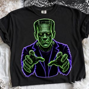 May include: A black short-sleeve t-shirt featuring a graphic print of a green-skinned monster with stitches on its forehead, wearing a dark purple jacket. The monster is depicted with its hands reaching forward in a glowing neon green outline.