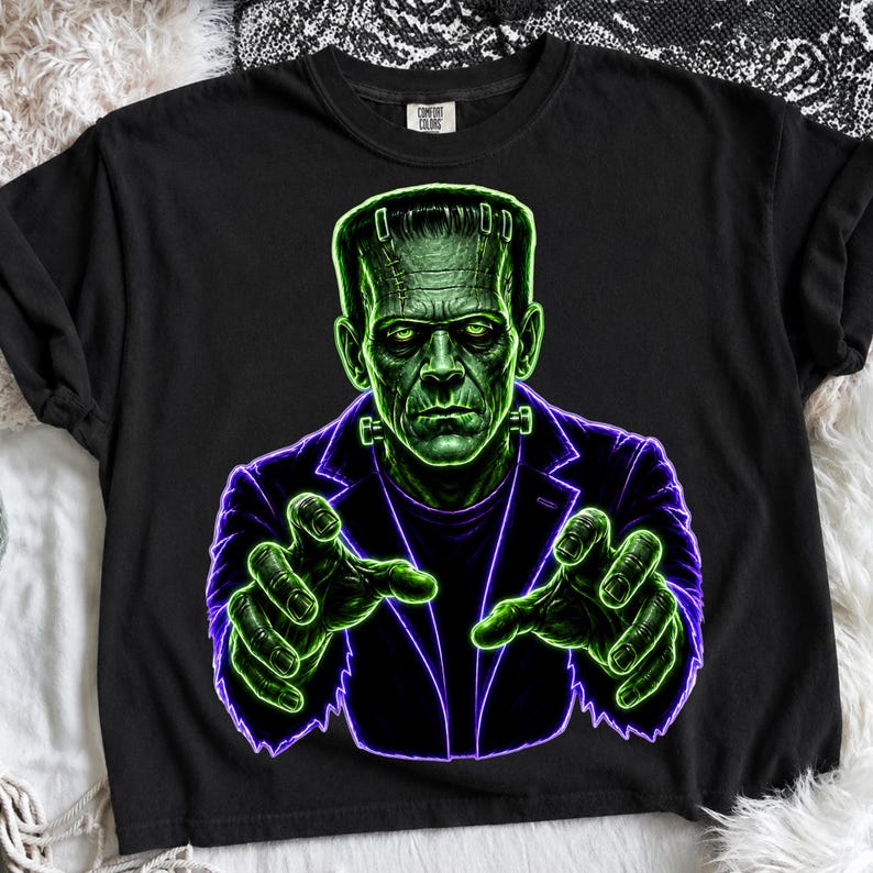 May include: A black short-sleeve t-shirt featuring a graphic print of a green-skinned monster with stitches on its forehead, wearing a dark purple jacket. The monster is depicted with its hands reaching forward in a glowing neon green outline.