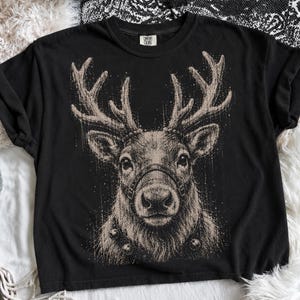 May include: A black short-sleeve t-shirt featuring a large, detailed graphic of a reindeer head with antlers. The reindeer is wearing a harness and a collar with bells. The graphic is rendered in a textured, sketch-like beige ink style.