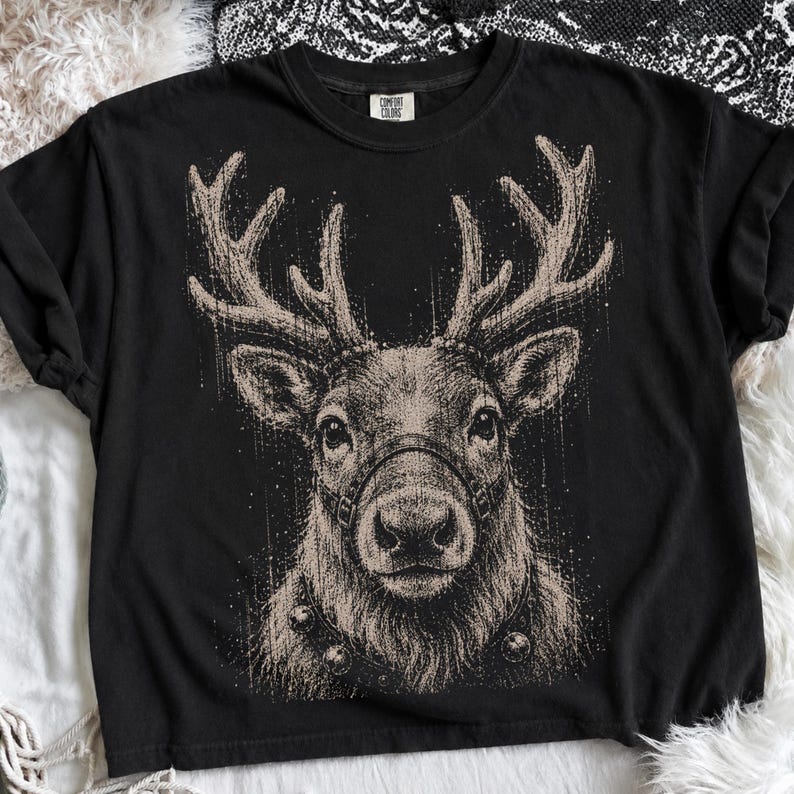 May include: A black short-sleeve t-shirt featuring a large, detailed graphic of a reindeer head with antlers. The reindeer is wearing a harness and a collar with bells. The graphic is rendered in a textured, sketch-like beige ink style.