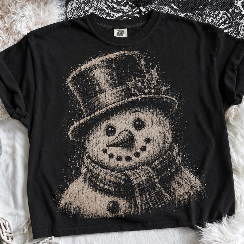 May include: A black short-sleeve t-shirt featuring a distressed beige graphic of a snowman wearing a top hat with holly leaves and a plaid scarf. The snowman has a carrot nose and a dotted smile. The shirt has a Comfort Colors brand label.