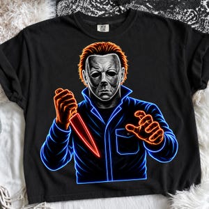 May include: A black t-shirt featuring a graphic of a masked figure with orange hair holding a knife. The design uses neon blue and orange outlines to create a glowing effect. The figure wears a dark jacket with a collar and a chest pocket.