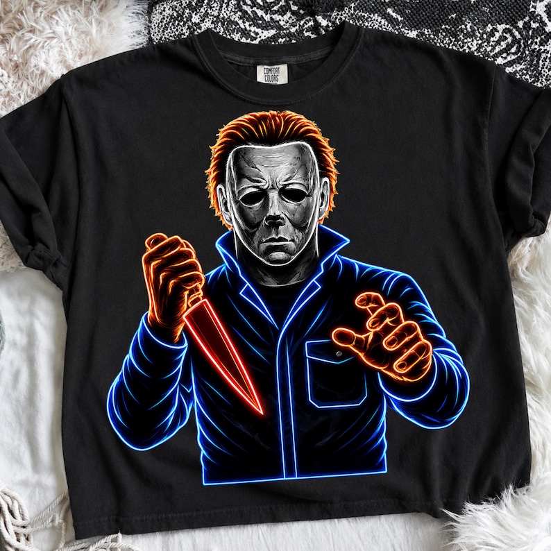 May include: A black t-shirt featuring a graphic of a masked figure with orange hair holding a knife. The design uses neon blue and orange outlines to create a glowing effect. The figure wears a dark jacket with a collar and a chest pocket.
