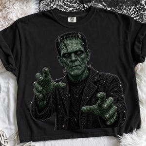 May include: A black short-sleeve t-shirt featuring a graphic of Frankenstein's monster rendered in a sparkling, rhinestone-like texture. The monster has green skin and wears a black jacket, with both hands reaching forward in a classic pose.