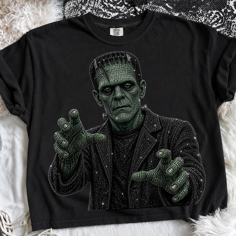 May include: A black short-sleeve t-shirt featuring a graphic of Frankenstein's monster rendered in a sparkling, rhinestone-like texture. The monster has green skin and wears a black jacket, with both hands reaching forward in a classic pose.
