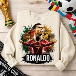 May include: A cream-colored crewneck sweatshirt featuring a graphic print of Cristiano Ronaldo. The design includes a portrait of the athlete, a back view of a player in a red number 7 jersey, the Portugal national crest, and the text RONALDO CR7.