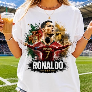 May include: A white t-shirt featuring a graphic print of soccer player Cristiano Ronaldo. The design includes a portrait, a back view of the player in a red jersey with the number 7, the Portugal national team crest, and the text RONALDO CR7.