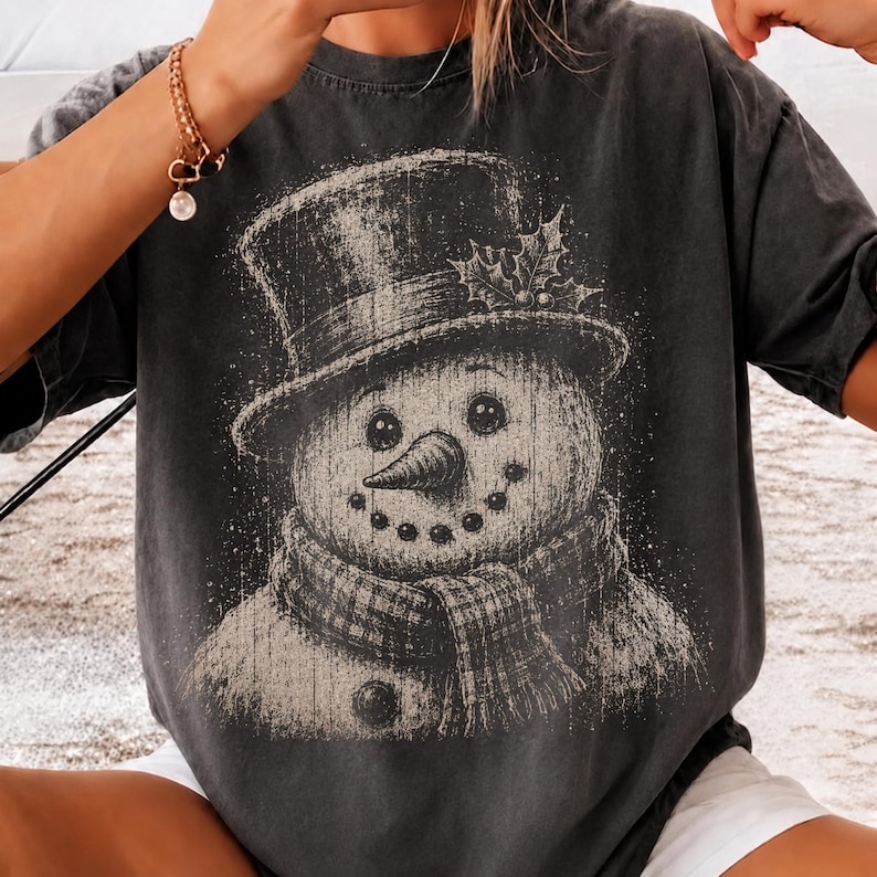 May include: A charcoal gray short-sleeve t-shirt featuring a distressed, vintage-style graphic of a snowman wearing a top hat with holly leaves and a plaid scarf. The snowman has a carrot nose and a dotted smile, rendered in a faded white ink.
