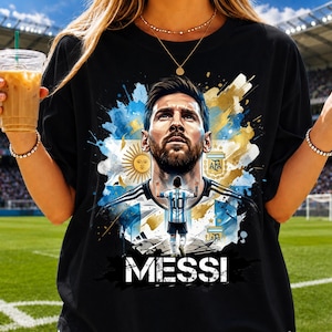 May include: A black t-shirt featuring a graphic print of Lionel Messi. The design includes a portrait of the athlete, a smaller figure wearing a number 10 jersey, the Argentina flag, the AFA logo, and the text MESSI in bold white letters.