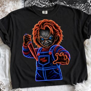 May include: A black t-shirt featuring a neon-style graphic of a doll with bright orange hair, glowing blue eyes, and a scarred face. The doll wears blue overalls with the text Good Guys printed on the chest and holds a sharp knife in one hand.