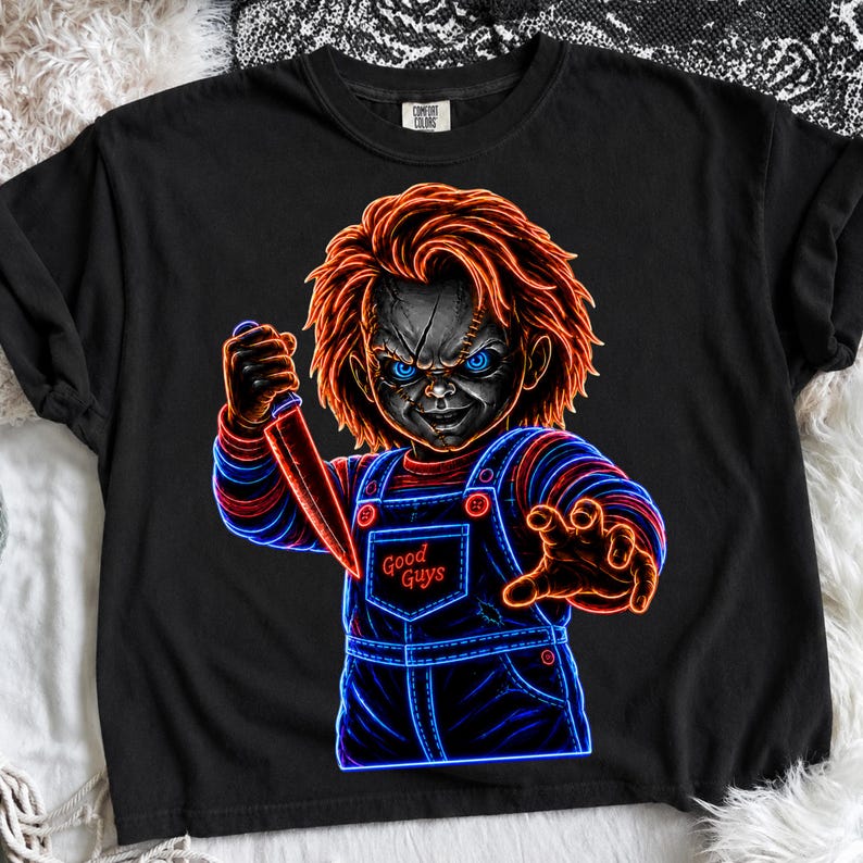 May include: A black t-shirt featuring a neon-style graphic of a doll with bright orange hair, glowing blue eyes, and a scarred face. The doll wears blue overalls with the text Good Guys printed on the chest and holds a sharp knife in one hand.