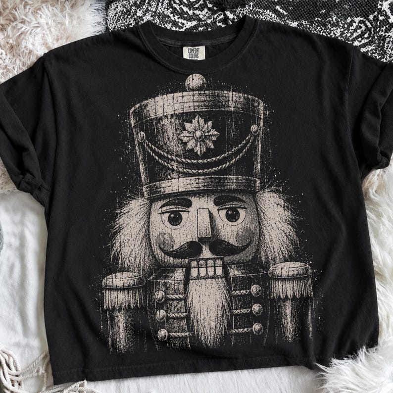 May include: A black short-sleeve t-shirt featuring a large, distressed-style graphic of a nutcracker soldier in shades of off-white and gray. The nutcracker wears a tall hat with a star emblem, has a mustache, and a long beard. The Comfort Colors label is visible.