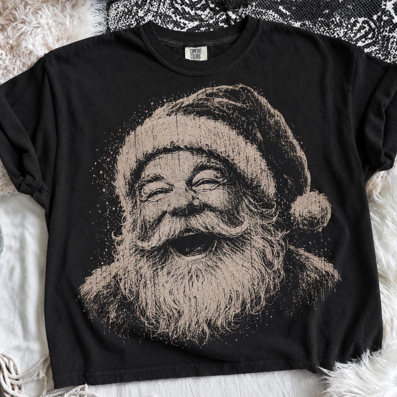 May include: A black short-sleeve t-shirt featuring a distressed, tan-colored graphic print of a laughing Santa Claus on the front. The graphic shows Santa with a full beard and a hat. The shirt has a Comfort Colors brand label at the inner neckline.