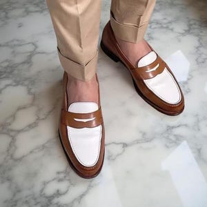 May include: A pair of two-tone penny loafers featuring a tan leather base with a white leather vamp and a classic strap across the top. The shoes are worn with cuffed tan trousers, showcasing a polished finish and a traditional slip-on design.