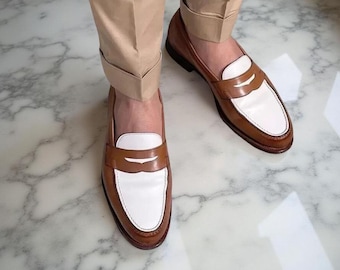 May include: A pair of two-tone penny loafers featuring a tan leather base with a white leather vamp and a classic strap across the top. The shoes are worn with cuffed tan trousers, showcasing a polished finish and a traditional slip-on design.