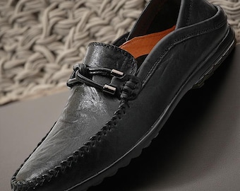 May include: A black slip-on loafer featuring a textured finish, a moccasin-style stitched toe seam, and a decorative strap across the vamp with two metallic silver accents. The interior lining is orange, and the shoe has a black rubber sole.