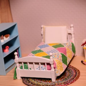 May include: A miniature dollhouse bedroom scene featuring a white bed with a colorful patchwork quilt, a blue bookshelf, and a small house. The bed sits on a round, multi-colored rug.