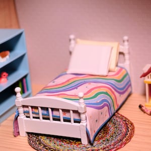 May include: A miniature bedroom scene with a white bed, rainbow quilt, and pillows. A light blue bookshelf holds toys and books. A small dollhouse and a colourful rug complete the scene.