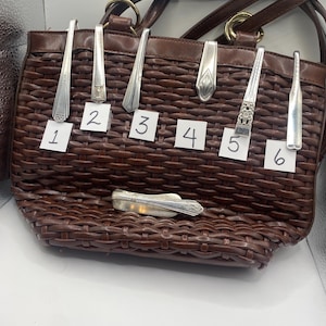 May include: A brown woven handbag with a leather handle and gold-coloured rings. Six silver spoons are inserted into the bag, each with a different design. A small silver knife is also visible.