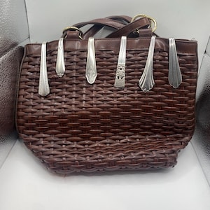 Peut inclure: Un sac à main marron tressé avec un dessus et des poignées en cuir. Des cuillères en argent sont attachées à l'avant du sac. Le sac a une forme rectangulaire et une texture tissée.