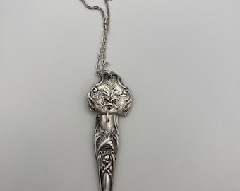 Puede incluir: Un collar con colgante plateado con un diseño floral ornamentado. El colgante, con forma de cuchara, presenta grabados detallados y un motivo floral. La delicada cadena plateada completa este elegante accesorio.