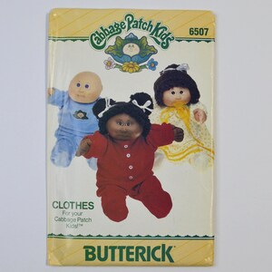 May include: A Butterick sewing pattern envelope for Cabbage Patch Kids clothes, number 6507. The package features illustrations of three dolls wearing a blue jumpsuit, a red button-front outfit, and a yellow patterned dress with white ribbons.