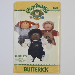 May include: A Butterick sewing pattern envelope 6508 for Cabbage Patch Kids clothes. The cover features three dolls wearing outfits including a blue striped shirt, a brown overall dress, and dark blue overalls with white shirts and sneakers.