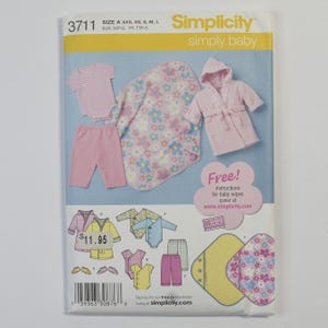 May include: A Simplicity 3711 sewing pattern envelope for baby clothing, featuring illustrations of a hooded robe, pants, a bodysuit, and a floral blanket. The package includes text for sizes XXS to L and instructions for baby wipes covers.