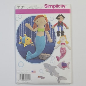 May include: A Simplicity 1131 sewing pattern envelope featuring illustrations of stuffed fabric toys including a mermaid with red hair, a pirate, a blonde mermaid with a pink patterned tail, a yellow and blue fish, and a gray shark.