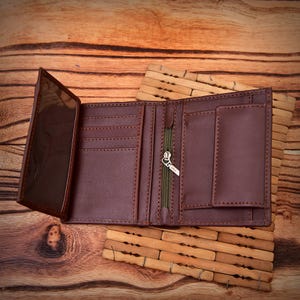 Puede incluir: Cartera de cuero marr&oacute;n abierta, mostrando m&uacute;ltiples ranuras para tarjetas, una ventana transparente para identificaci&oacute;n y un compartimento con cremallera. Se exhibe abierta sobre una superficie de madera, mostrando su interior.