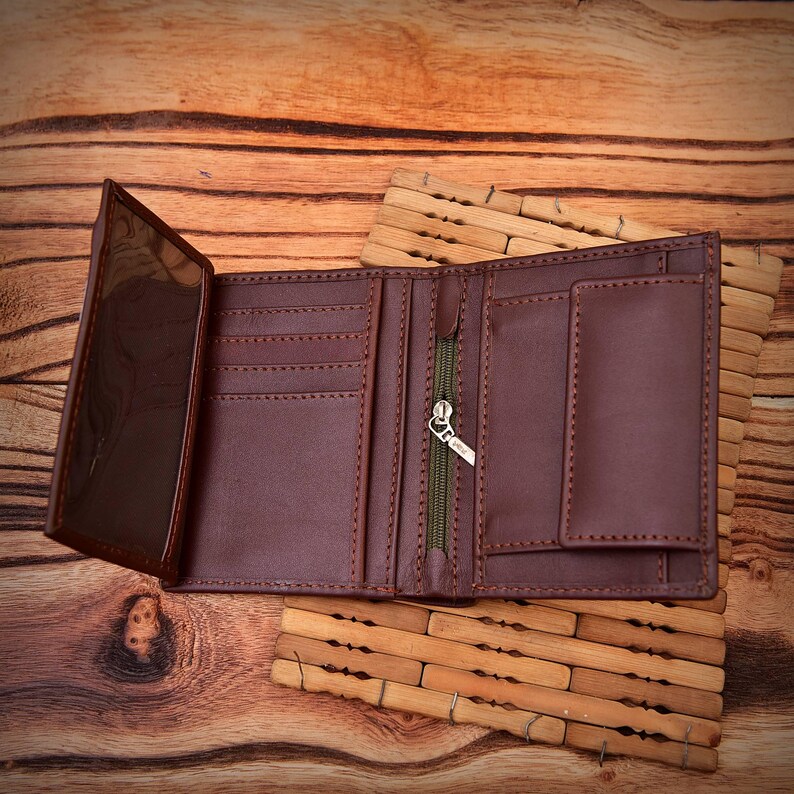 Puede incluir: Cartera de cuero marr&oacute;n abierta, mostrando m&uacute;ltiples ranuras para tarjetas, una ventana transparente para identificaci&oacute;n y un compartimento con cremallera. Se exhibe abierta sobre una superficie de madera, mostrando su interior.