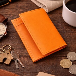 Puede incluir: Dos carteras de cuero naranja apiladas sobre una superficie de madera, con un llavero, llaves y monedas cerca. Tambi&eacute;n se ven un bol&iacute;grafo, gafas y una taza de caf&eacute;. Las carteras tienen un borde cosido.