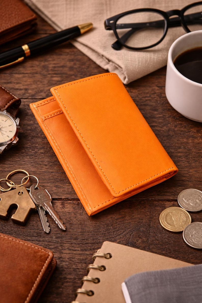 Puede incluir: Dos carteras de cuero naranja apiladas sobre una superficie de madera, con un llavero, llaves y monedas cerca. Tambi&eacute;n se ven un bol&iacute;grafo, gafas y una taza de caf&eacute;. Las carteras tienen un borde cosido.