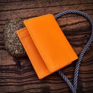 Puede incluir: Cartera de cuero naranja brillante con costuras visibles. La cartera est&aacute; abierta, revelando ranuras para tarjetas. Una piedra gris y una cuerda azul en el fondo. Sobre una superficie de madera oscura.