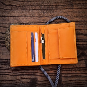 Puede incluir: Cartera de cuero naranja brillante abierta, con m&uacute;ltiples ranuras para tarjetas, un compartimento con cremallera y un bolsillo peque&ntilde;o. La cartera est&aacute; cosida con un hilo en contraste y tiene un tirador de cremallera plateado. Se muestra sobre una superficie de madera.