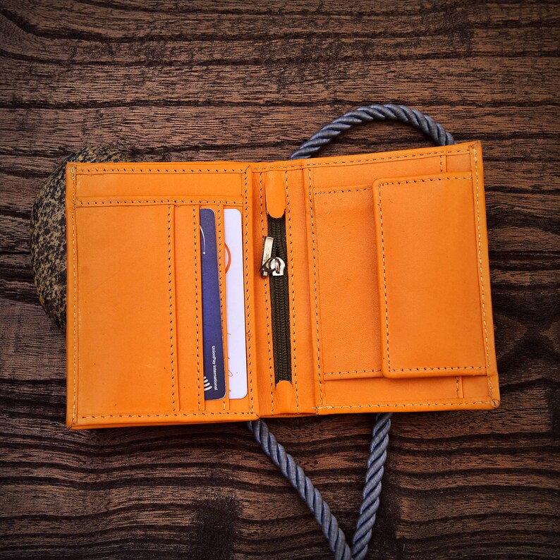 Puede incluir: Cartera de cuero naranja brillante abierta, con m&uacute;ltiples ranuras para tarjetas, un compartimento con cremallera y un bolsillo peque&ntilde;o. La cartera est&aacute; cosida con un hilo en contraste y tiene un tirador de cremallera plateado. Se muestra sobre una superficie de madera.