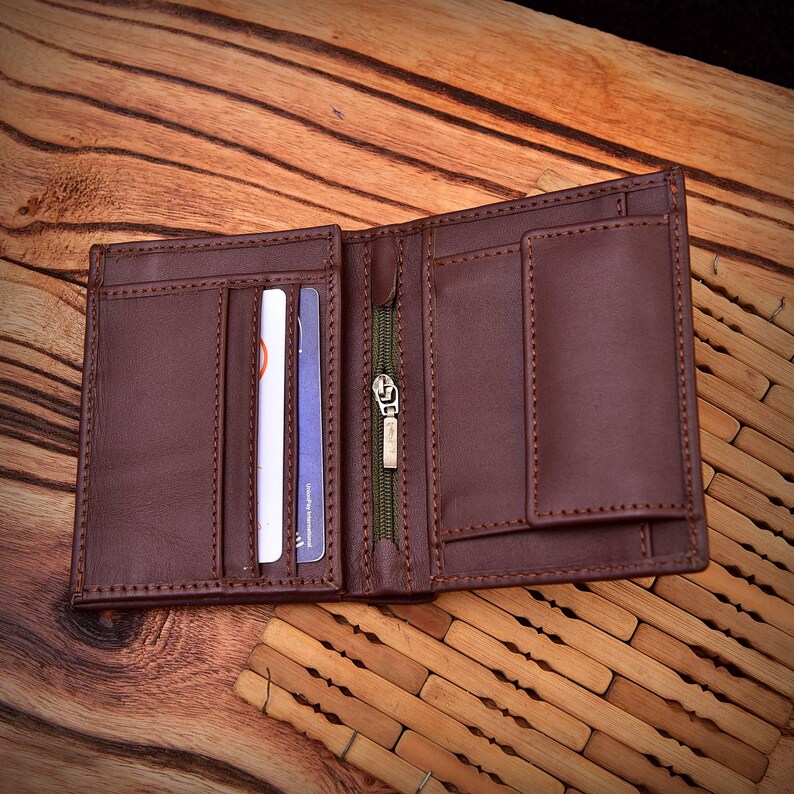 Puede incluir: Cartera de cuero marr&oacute;n abierta con m&uacute;ltiples ranuras para tarjetas, un compartimento con cremallera y un billetero. La cartera est&aacute; hecha de cuero cosido y se muestra abierta, mostrando su dise&ntilde;o interior. El fondo es una superficie de madera.