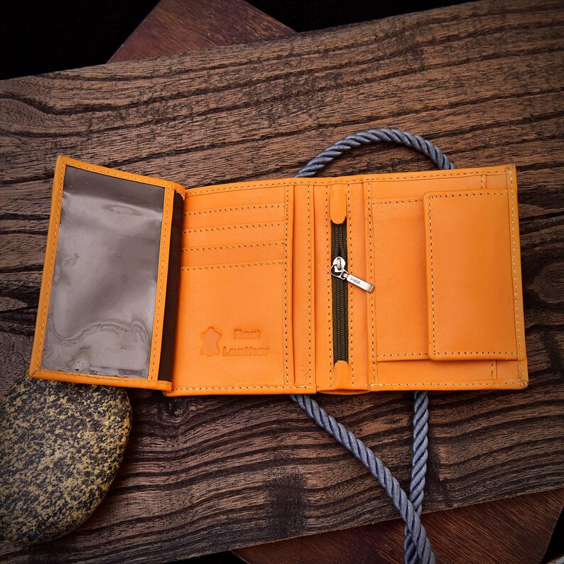 Puede incluir: Cartera de cuero naranja abierta, con m&uacute;ltiples compartimentos, incluyendo una ventana de identificaci&oacute;n transparente y un bolsillo con cremallera. La cartera se muestra abierta, mostrando su dise&ntilde;o interior y construcci&oacute;n. La cartera est&aacute; sobre una superficie de madera.