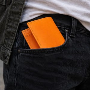Puede incluir: Cartera de cuero naranja brillante en un bolsillo de mezclilla oscura. La cartera es rectangular con costuras visibles. El bolsillo es parte de unos jeans de mezclilla negros. Tambi&eacute;n se ve una camiseta blanca y una chaqueta de mezclilla negra.