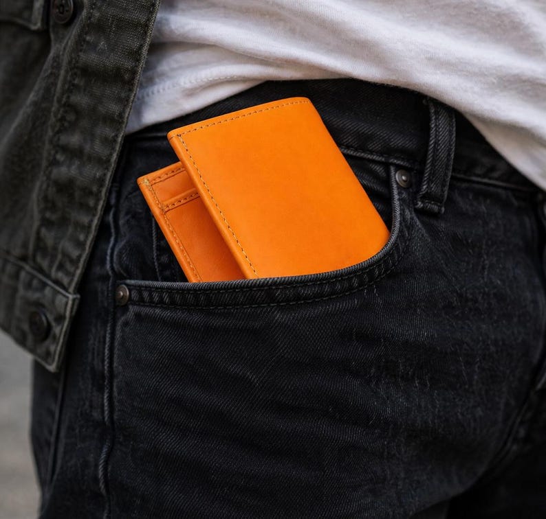 Puede incluir: Cartera de cuero naranja brillante en un bolsillo de mezclilla oscura. La cartera es rectangular con costuras visibles. El bolsillo es parte de unos jeans de mezclilla negros. Tambi&eacute;n se ve una camiseta blanca y una chaqueta de mezclilla negra.