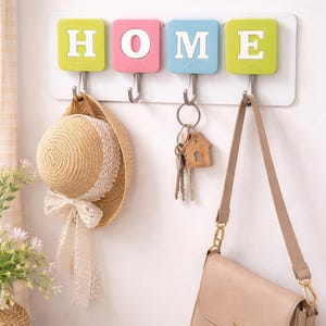 Puede incluir: Un llavero de pared con la palabra "HOME" escrita en baldosas cuadradas de colores. Un sombrero de paja, llaves y un bolso cuelgan de los ganchos de abajo. El sombrero tiene una cinta de encaje blanco. El bolso es beige.