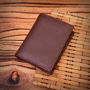 Puede incluir: Una cartera de cuero marr&oacute;n con bordes cosidos. La cartera rectangular est&aacute; cerrada. El fondo es una superficie de madera y una esterilla de bamb&uacute;.